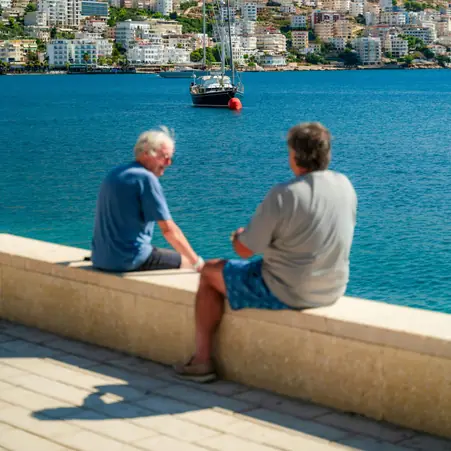 Two older men talking on a seafront wall, with a bay and a sailing boat behind them